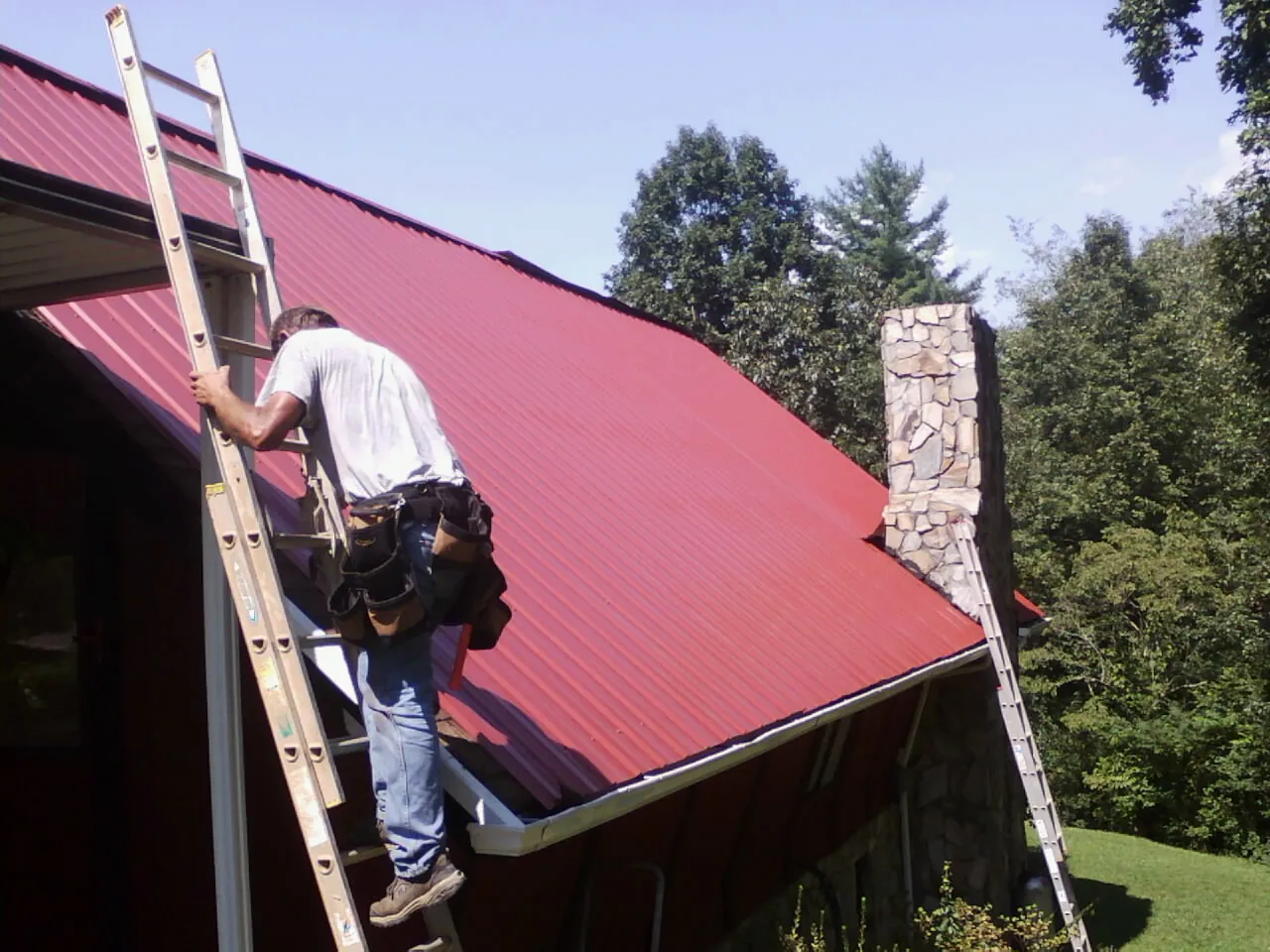 Roofer on a ladder beside a red metal roof and stone chimney — Soffit & Fascia Repair by Roofing Brothers in Saint Peters